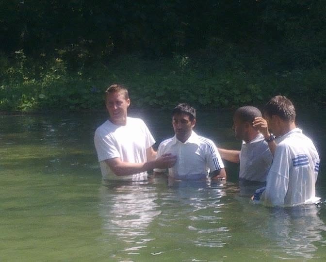 A group of men standing waist-deep in a river during a baptism, surrounded by trees